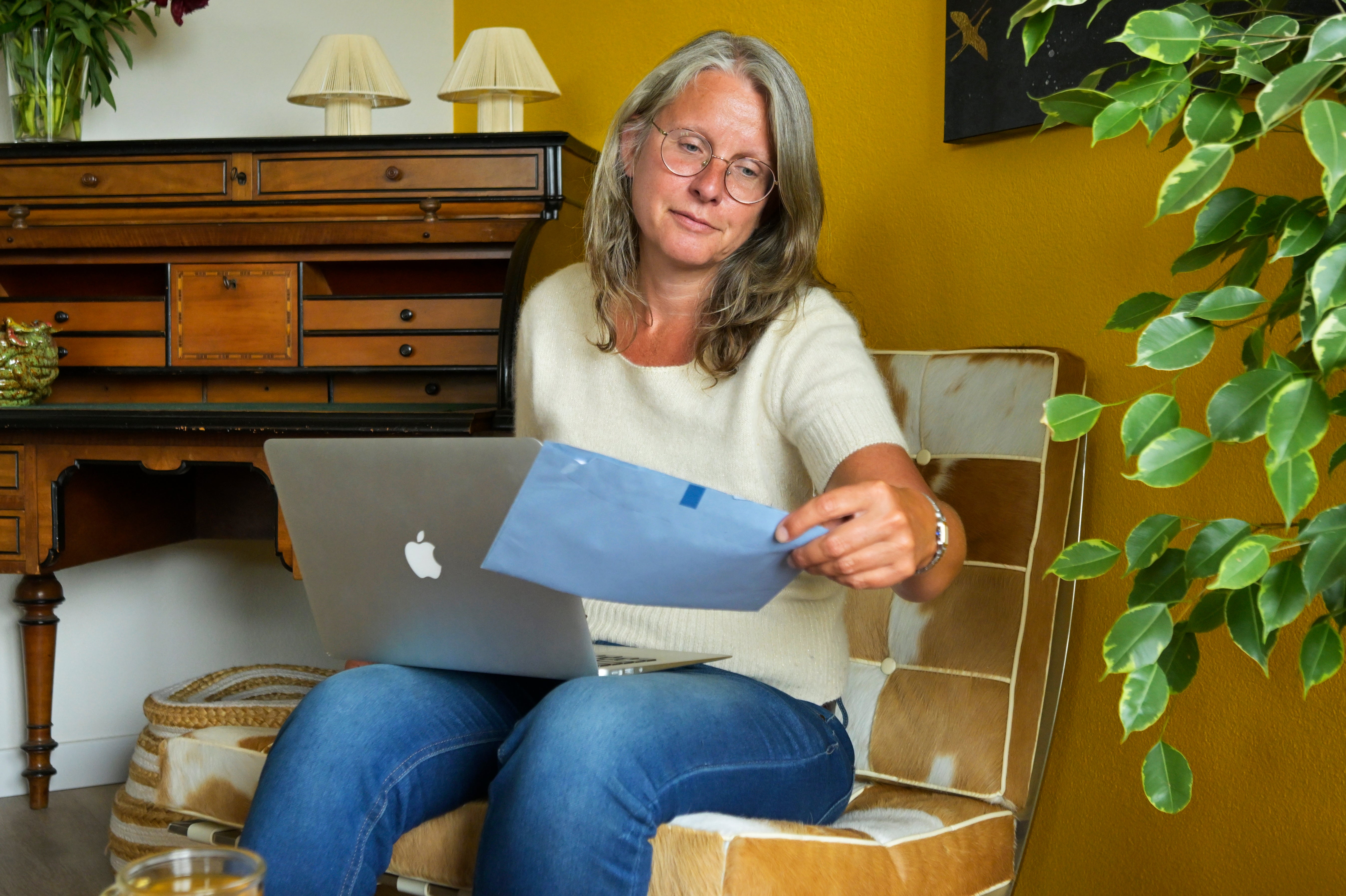 Vrouw op stoel in woonkamer met laptop op schoot bekijkt blauwe envelop van de Belastingdienst