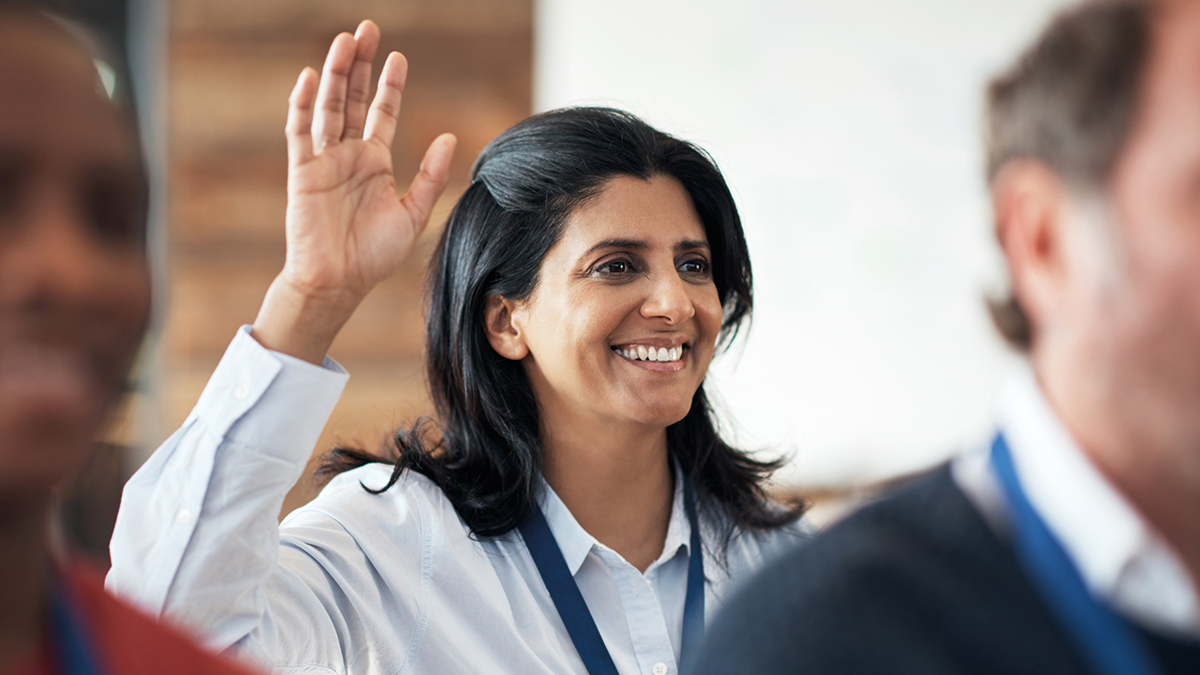 A person holding her right hand up, smiling, in a light blue shirt and lanyard. 