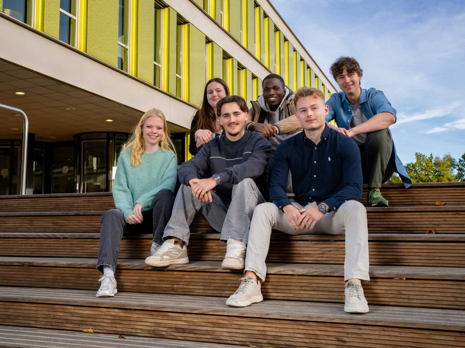 Groep Avansstudenten zitten buiten op een houten trap bij het Avansgebouw in Tilburg