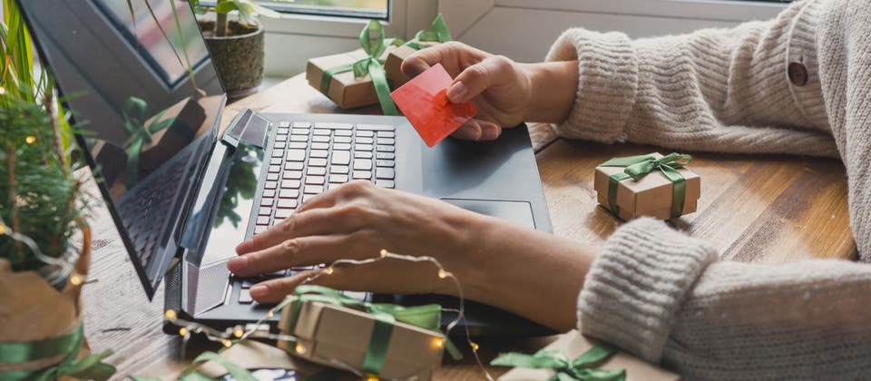 Close-up of hands using a laptop for online shopping, holding a red credit card. The desk is decorated with small gift boxes tied with green ribbons and festive lights, suggesting holiday shopping.