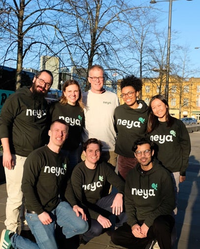 A group of people standing and kneeling outdoors, wearing black and white ‘neya’ branded hoodies and tops. The photo is taken in a paved outdoor area with buildings and trees visible in the background.