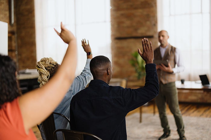 Several attendees seated in a workshop or training session raise their hands while a presenter stands at the front of a bright room with large windows and exposed brick walls.