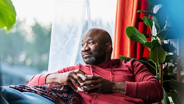 A man sitting on his sofa at home with a cup of coffee, looking out of a window, looking thoughtful.