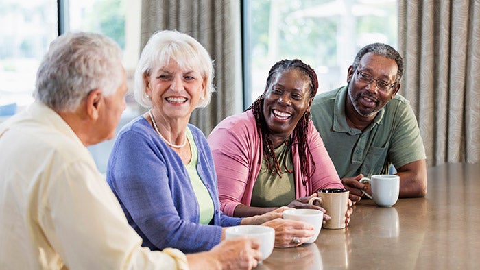 Four multi-ethnic mature and senior adults sitting indoors at a table, drinking coffee, conversing and smiling. The main focus is on an African-American couple in their 50s. Their friends are a senior couple in their 70s.
