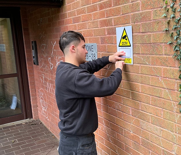 A man fixing a CCTV warning sign, to a red brick wall