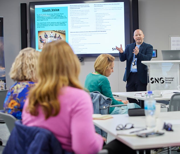 A man standing in front of a projector screen, talking to a group of people
