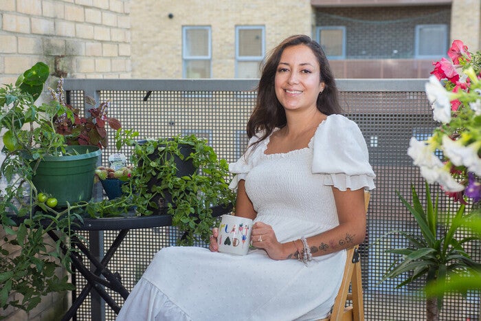 A lady wearing a white dress sitting on her balcony with a cup of tea, surrounded by pretty flowers