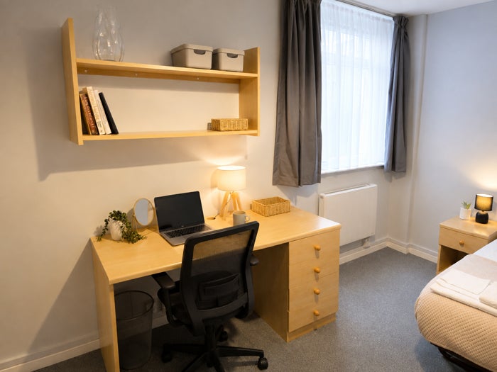 Furnished single bedroom with workspace, shelving and window.