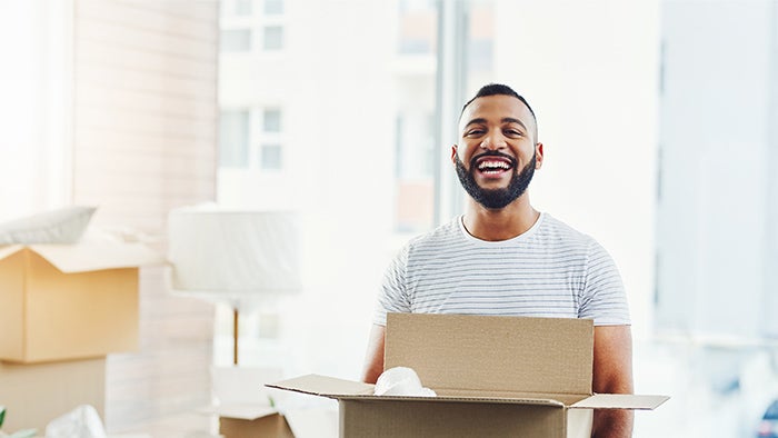 Person standing indoors holding an open cardboard box, with moving boxes visible in a bright, modern room