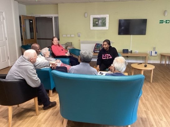Photo of six older people sitting in chairs listening to a facilitator for the Reminiscence programme.