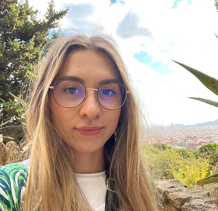 Close-up of a lady with long, blonde hair and glasses, smiling at the camera with a town in the background