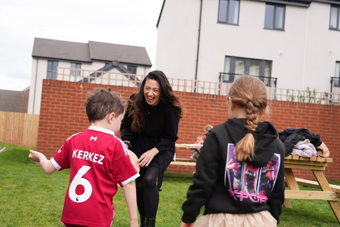 Three people interacting on a grassy area outside residential buildings, with picnic tables visible nearby