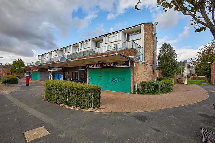 Local shopping parade with ground-floor retail units, flats above, paved walkways and communal outdoor space.