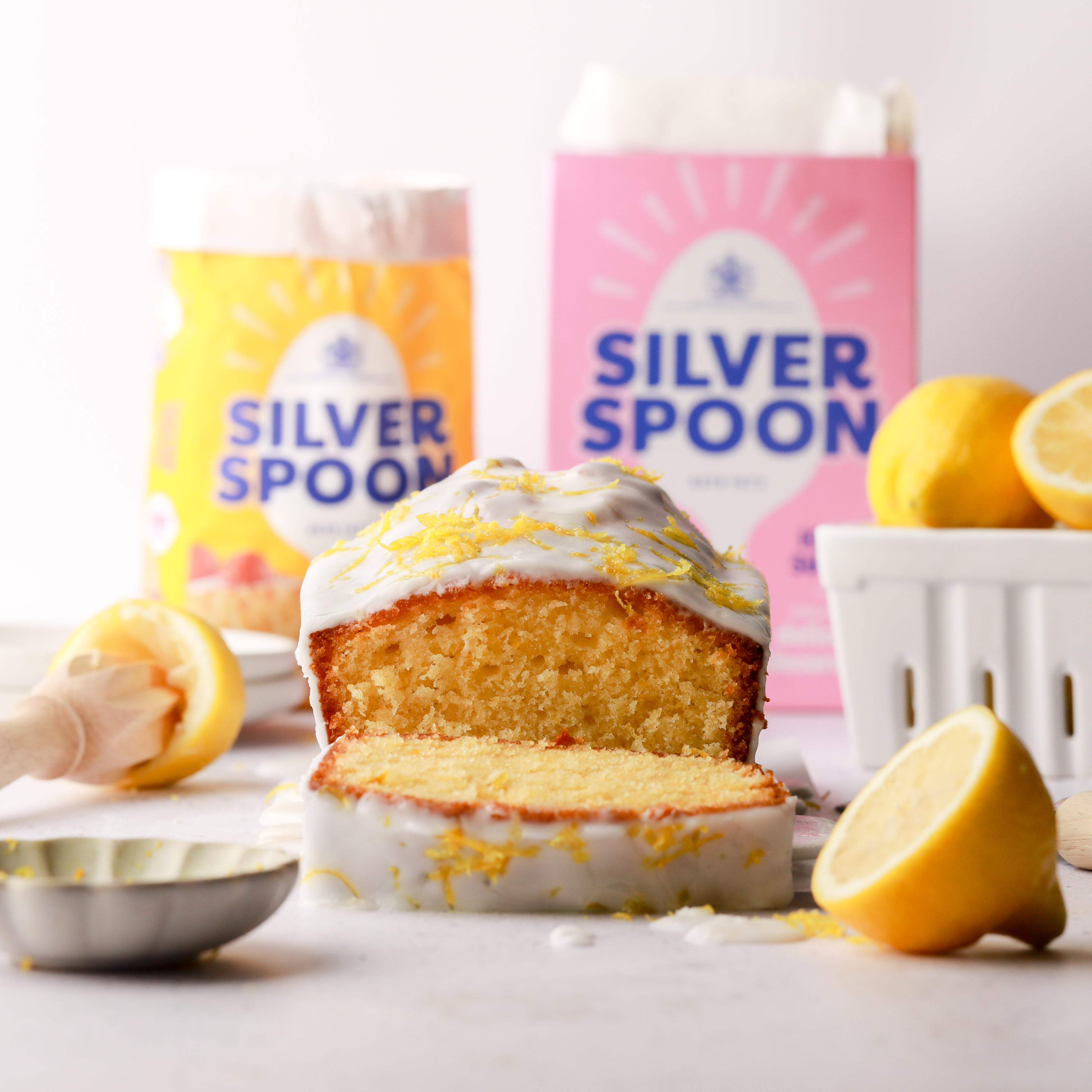 A freshly baked lemon drizzle cake with a slice cut off, with the pink and yellow packs of Silver Spoon icing and caster sugar in the background, amongst a messy kitchen scene.
