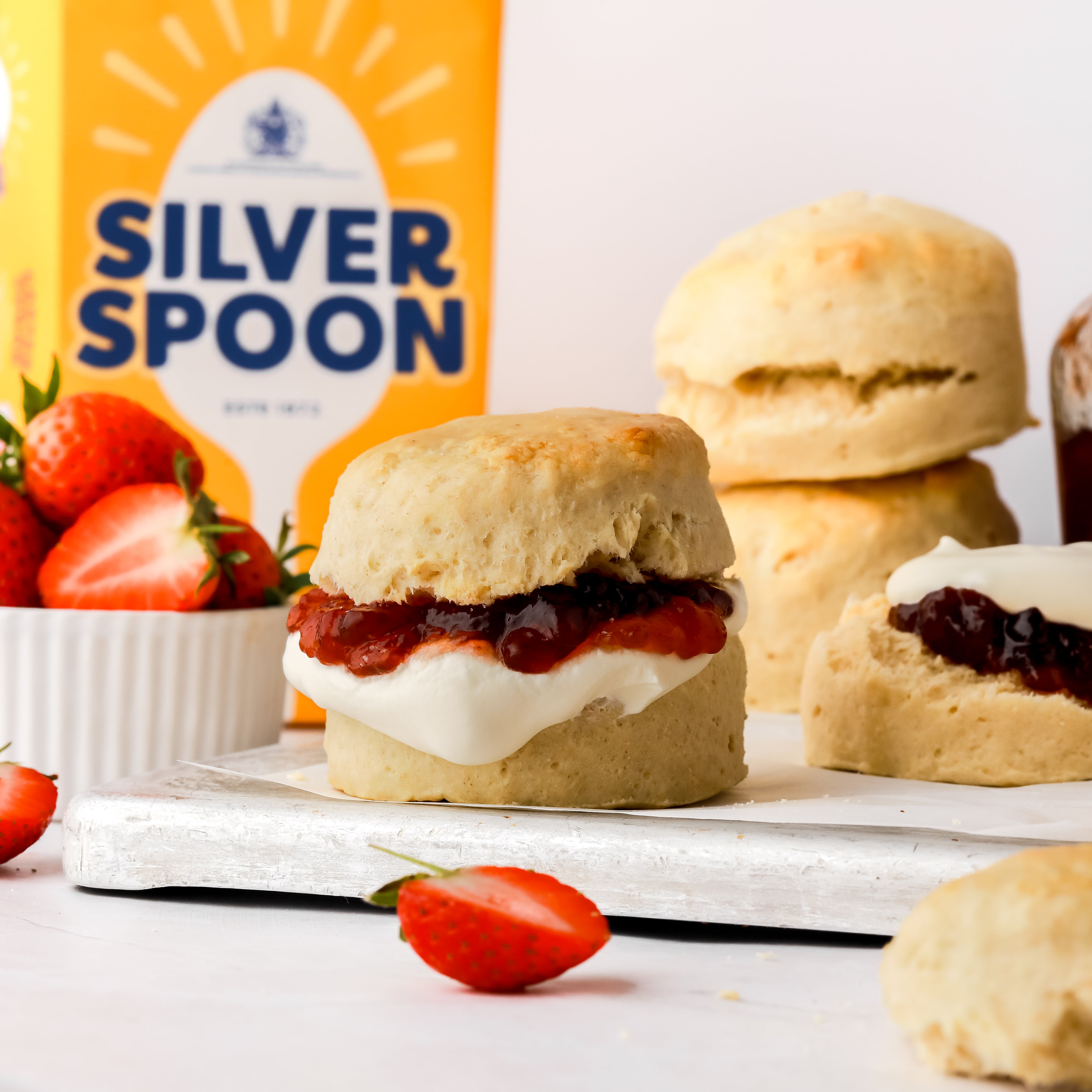 Freshly made scones with a small pot of sliced strawberries in front of a yellow bag of Silver Spoon caster sugar.