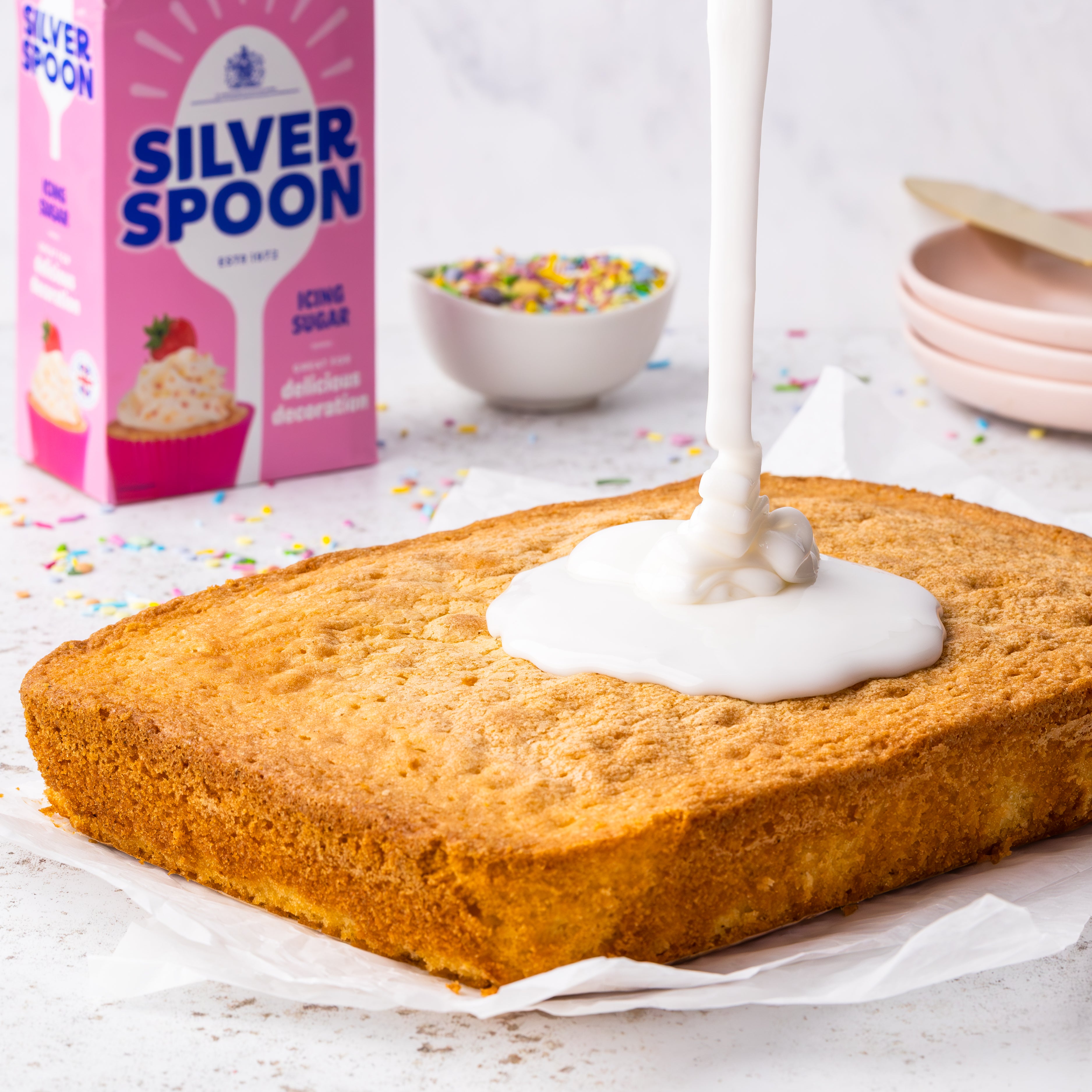 A freshly made tray-bake cake with icing being drizzled on top, in front of a pink pack of Silver Spoon icing sugar.