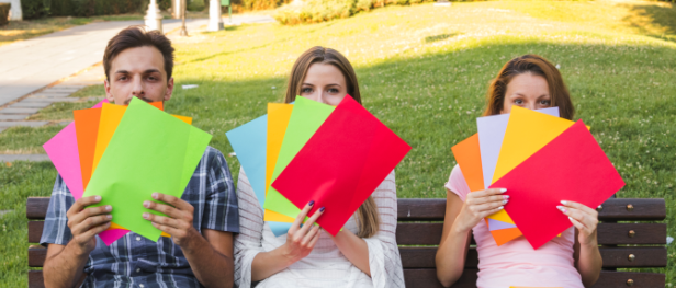 Three young people sit on a bench outside holding up coloured papers.
