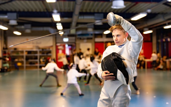Sverre Jansen is gekleed in de beschermende kleding die bij de sport schermen hoort. Hij houdt een floret omhoog en heeft een masker in zijn andere hand.