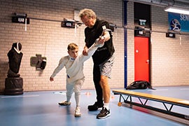 De jonge Sven in schermuitrusting krijgt schermles van een coach in een sporthal, terwijl ze samen een beweging oefenen