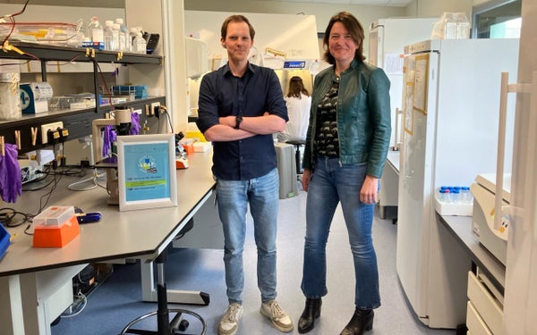 Twee onderzoekers staan in een laboratorium vol apparatuur en chemicaliën, glimlachend naar de camera. Een bord met "LEAF" is op de voorgrond te zien.