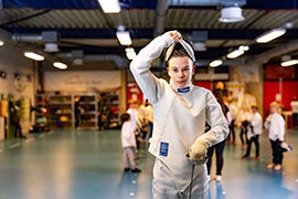 De jonge schermer Sven, gekleed in volledige uitrusting, zet zijn masker op in een sporthal, met andere kinderen op de achtergrond