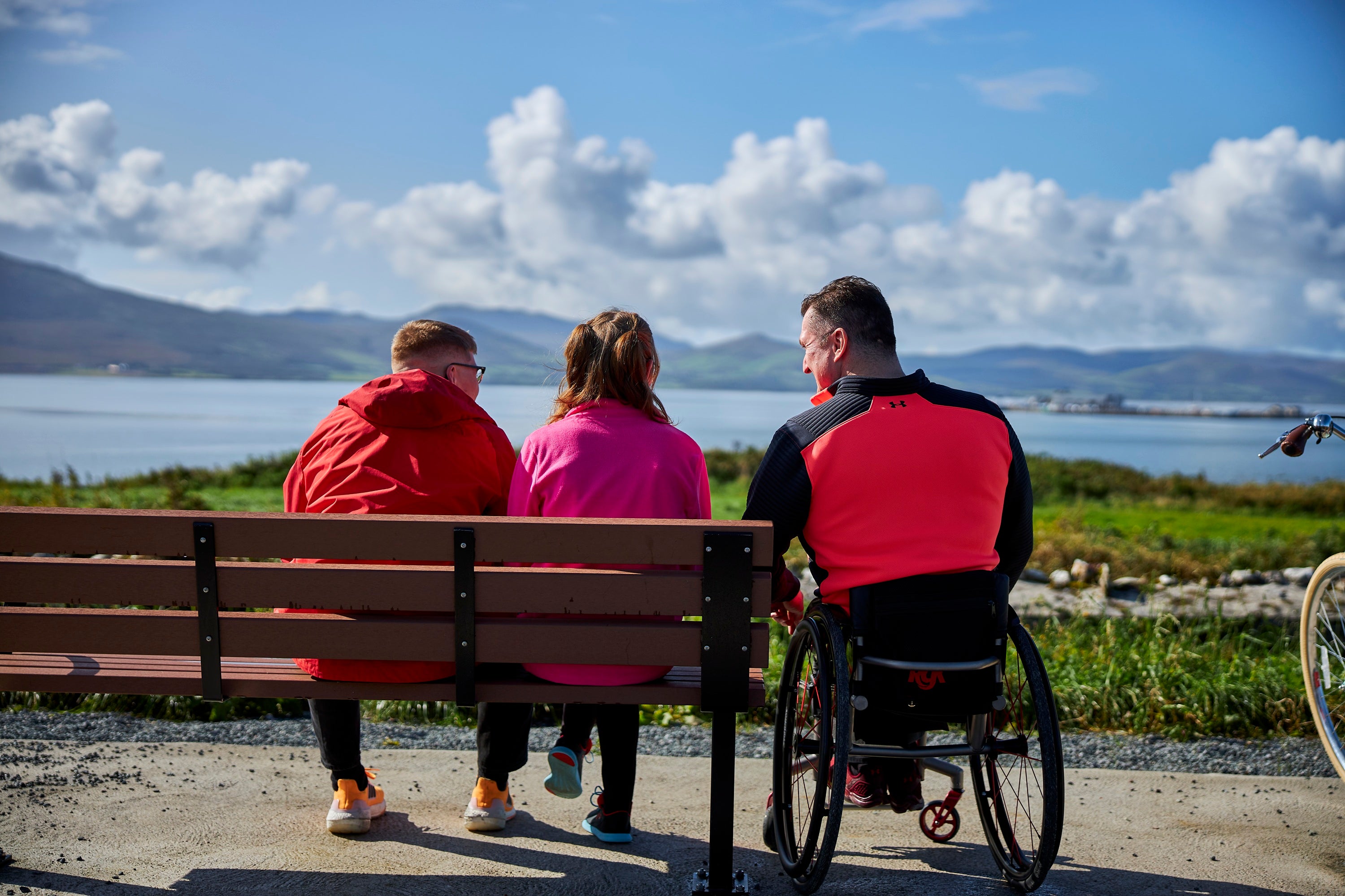 People sitting on a bench and one person in a wheelchair with coastal view