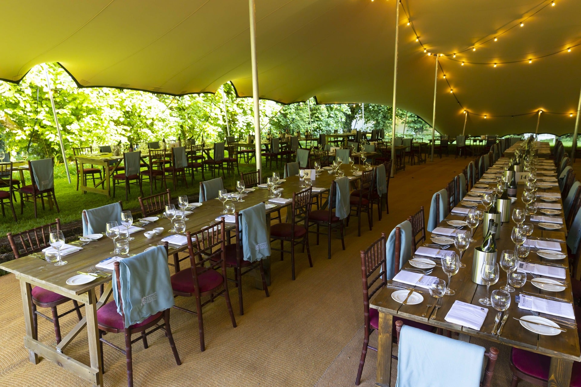 Interior of a large outdoor marquee set for a dinner event with long wooden tables, wine glasses and festoon lighting