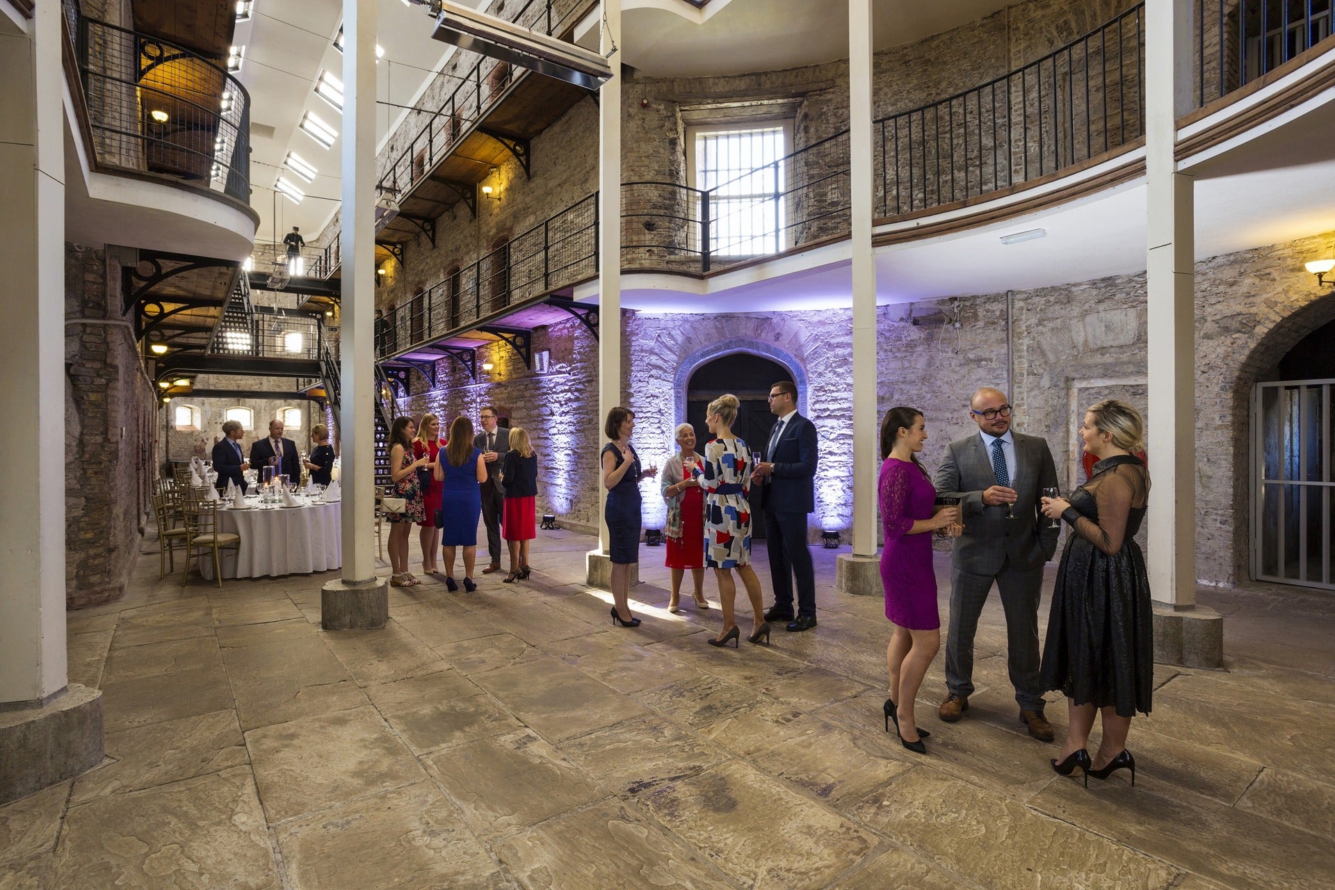 Guests networking at a formal drinks reception in a historic stone-walled venue Cork Jail with iron balconies and atmospheric lighting