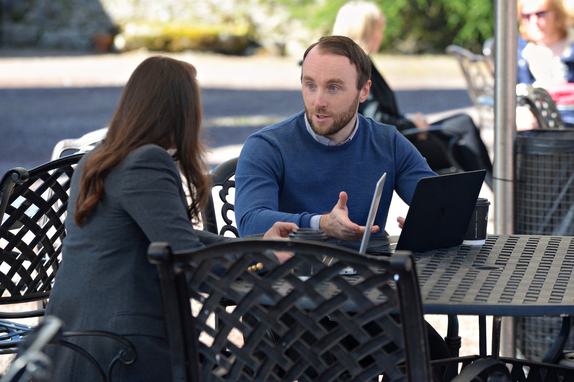 Two people having an outdoor meeting at a table with a laptop and takeaway coffee