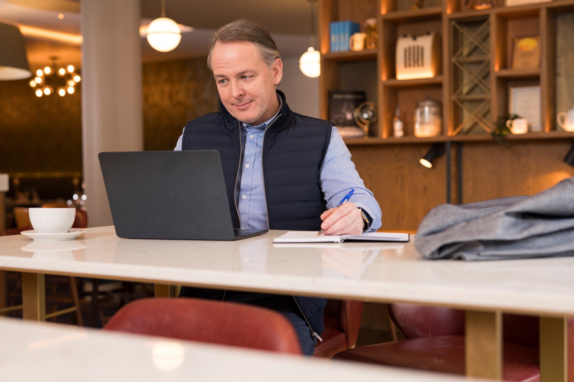 Man in a navy gilet working at a laptop and taking notes in a hotel lounge