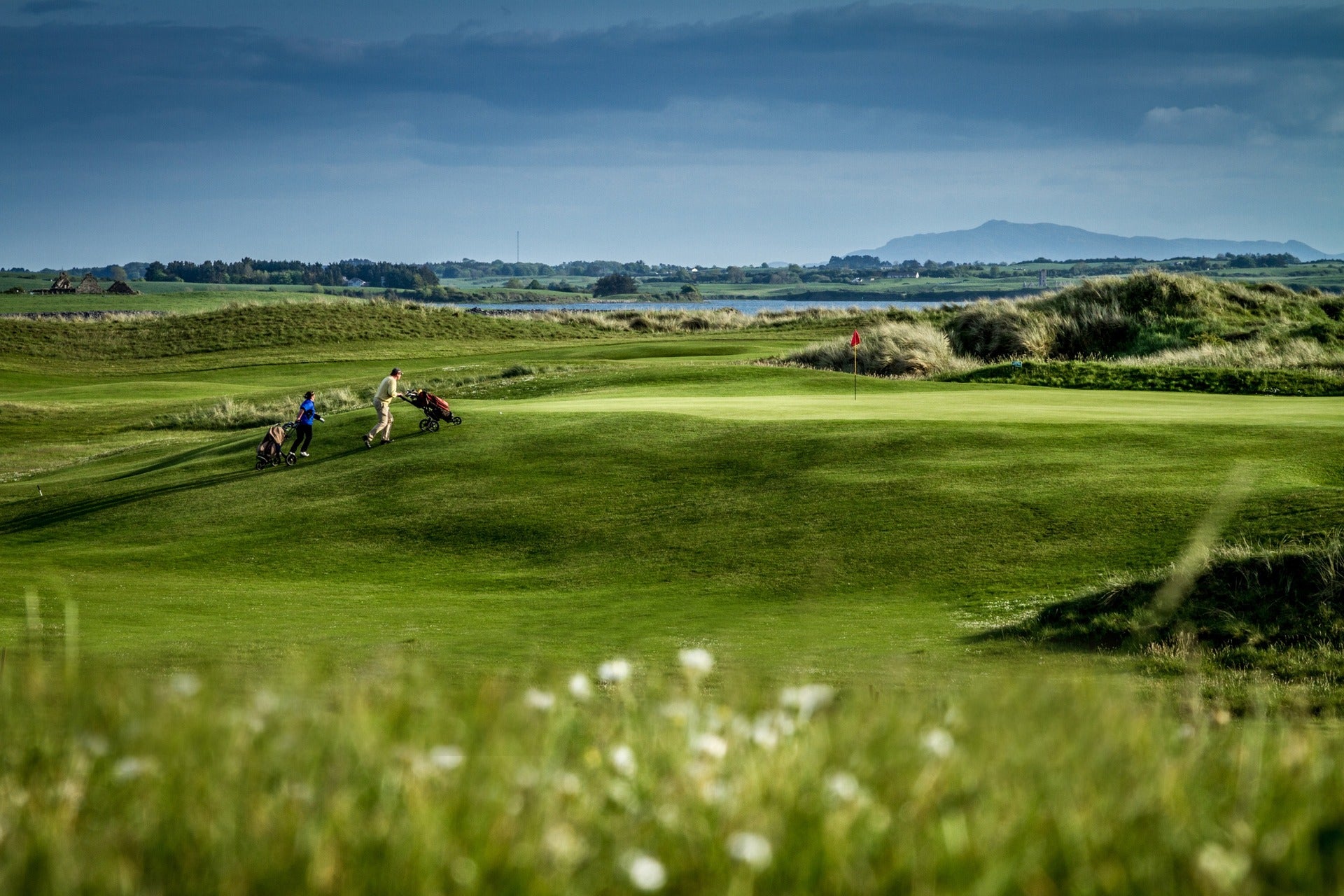Two golfers pushing trolleys along a links fairway with a coastal lake and mountains in the distance