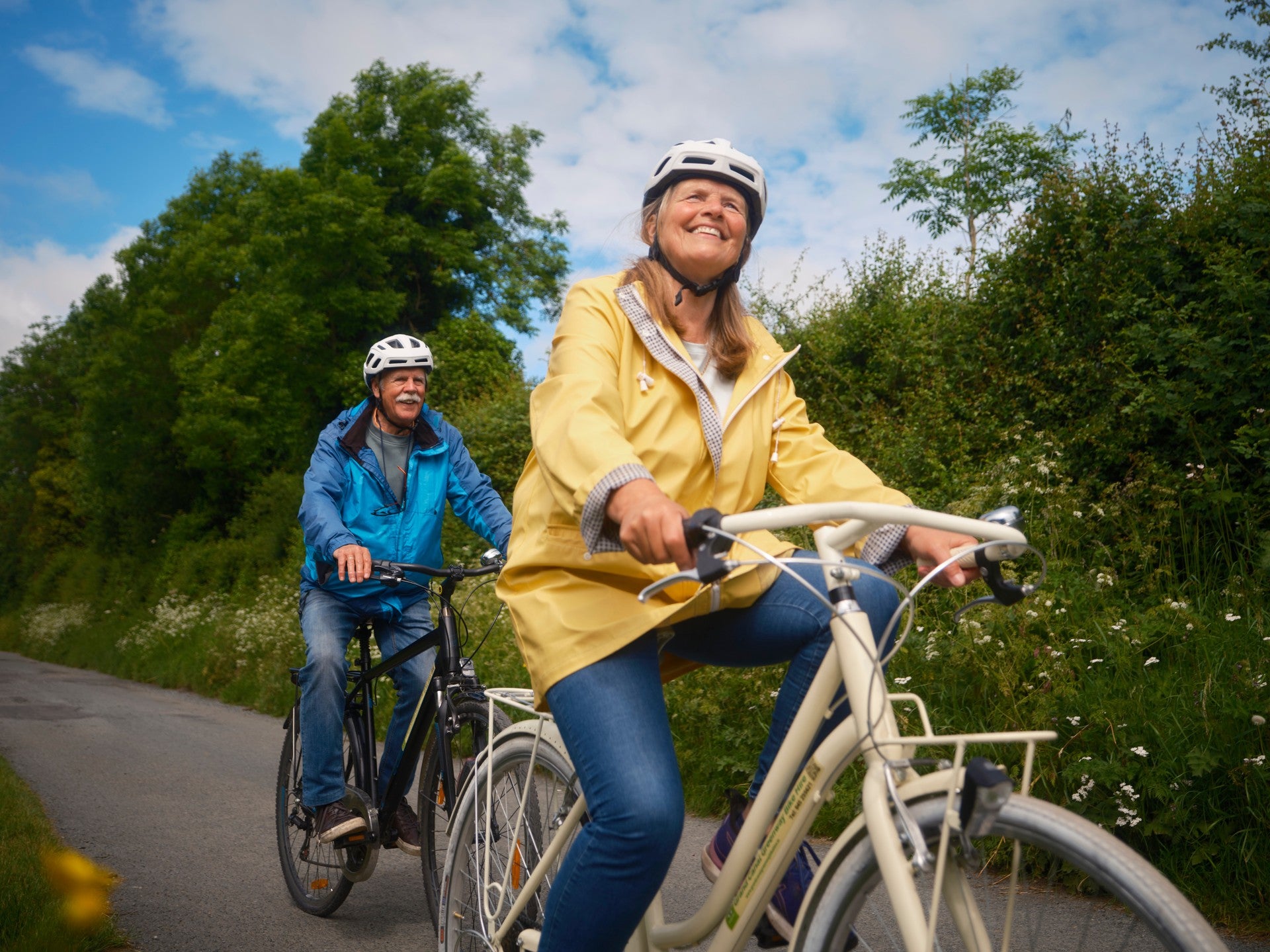 Two cyclists riding along a tree-lined greenway on a sunny day, both wearing helmets
