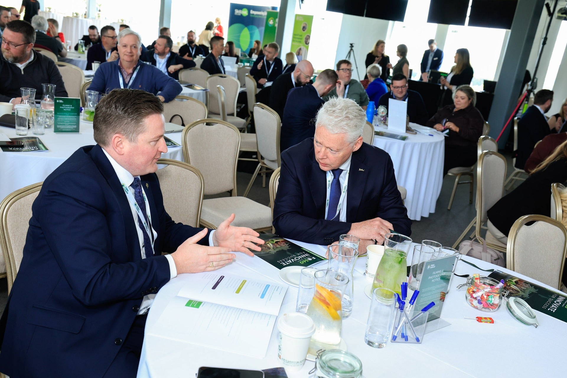 Two men in suits having a one-to-one business conversation at a conference table with other attendees in the background