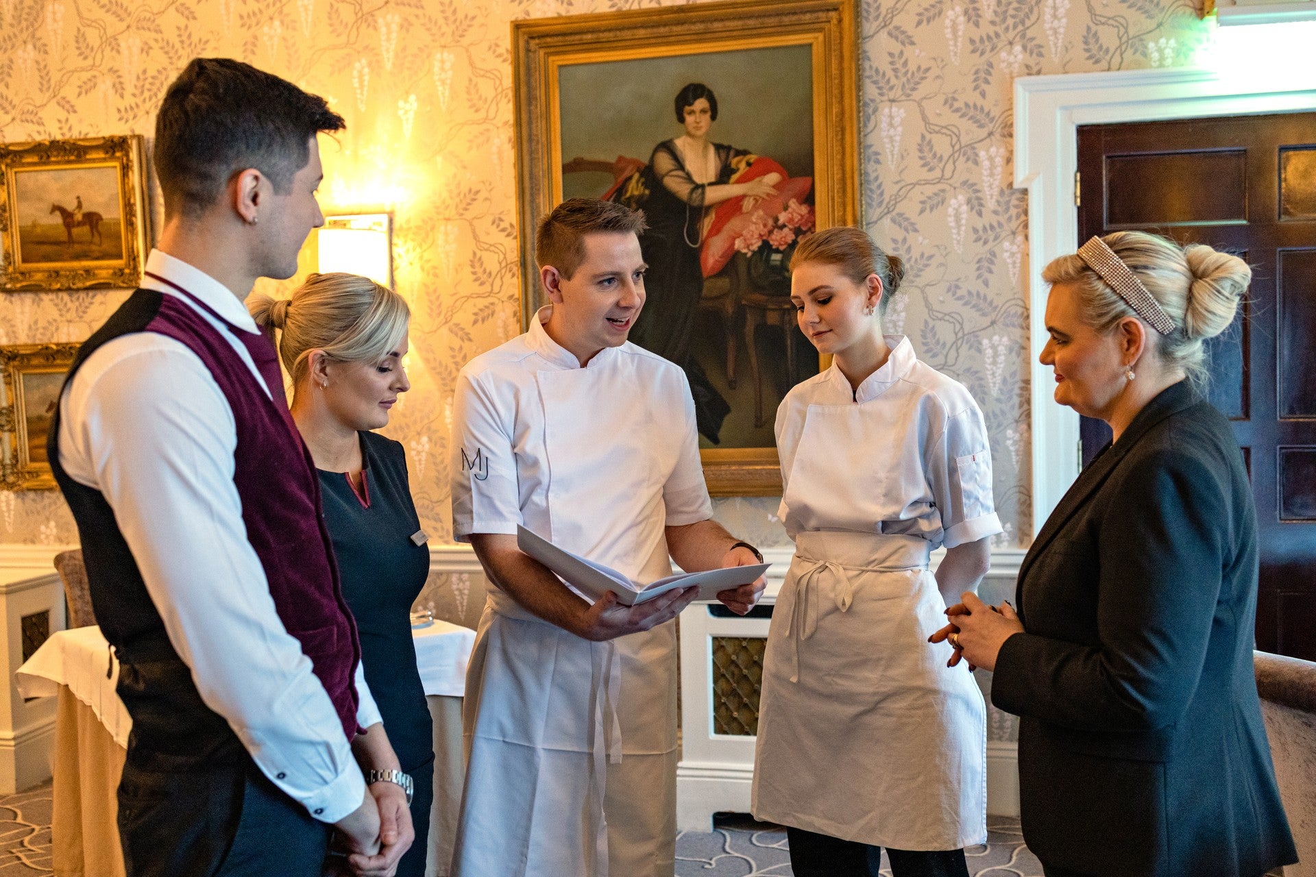 Five hospitality staff members including a chef in whites gathered in discussion in an elegantly decorated hotel room