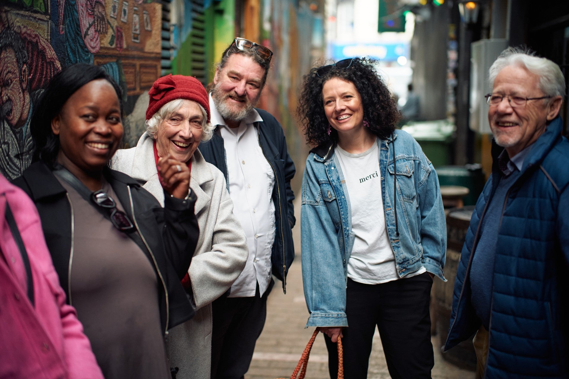 A group of five people of mixed ages laughing together in a colourful street art laneway