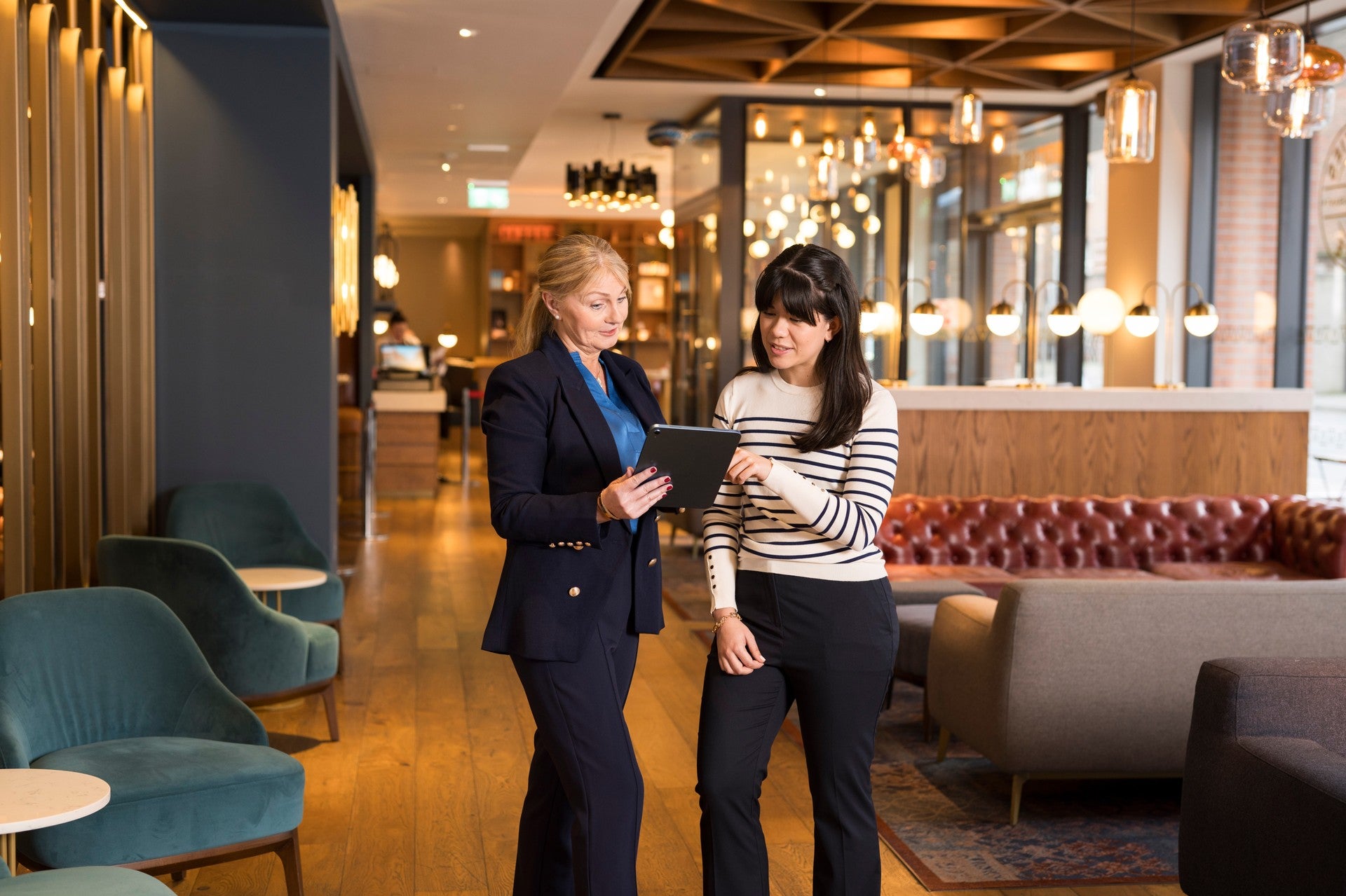 Two women reviewing content together on a tablet while standing in a hotel lobby lounge
