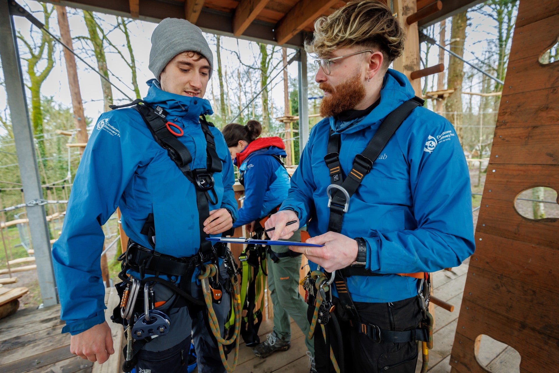 Two outdoor activity staff in harnesses checking safety equipment at a high ropes platform
