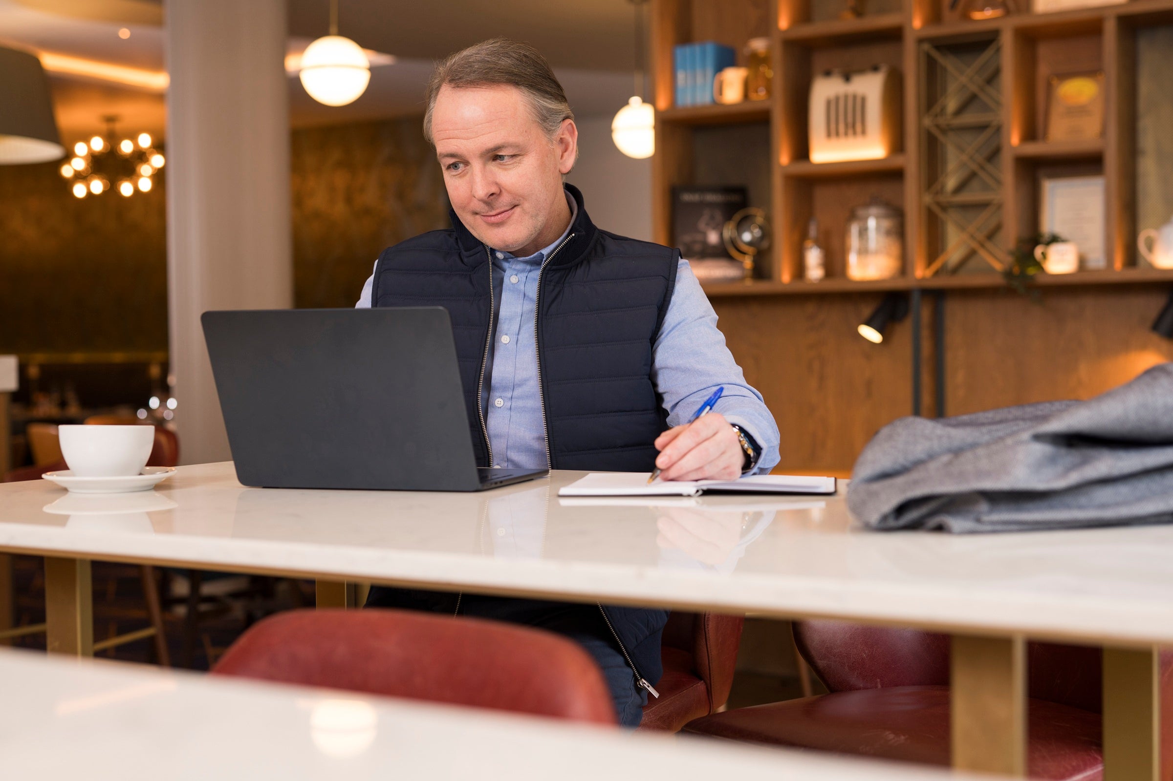 Smartly dressed man working on a laptop, in a hotel lobby, with a coffee.