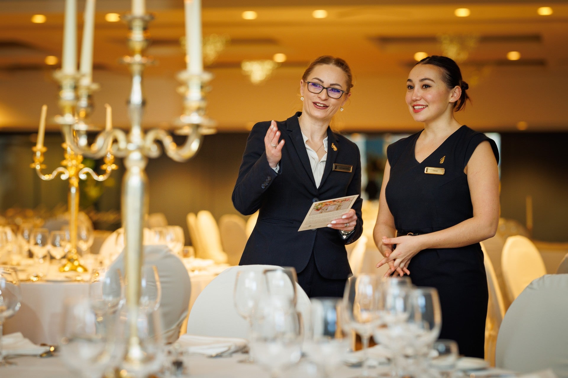 Two hotel staff members in formal uniforms speaking together in a set banquet dining room