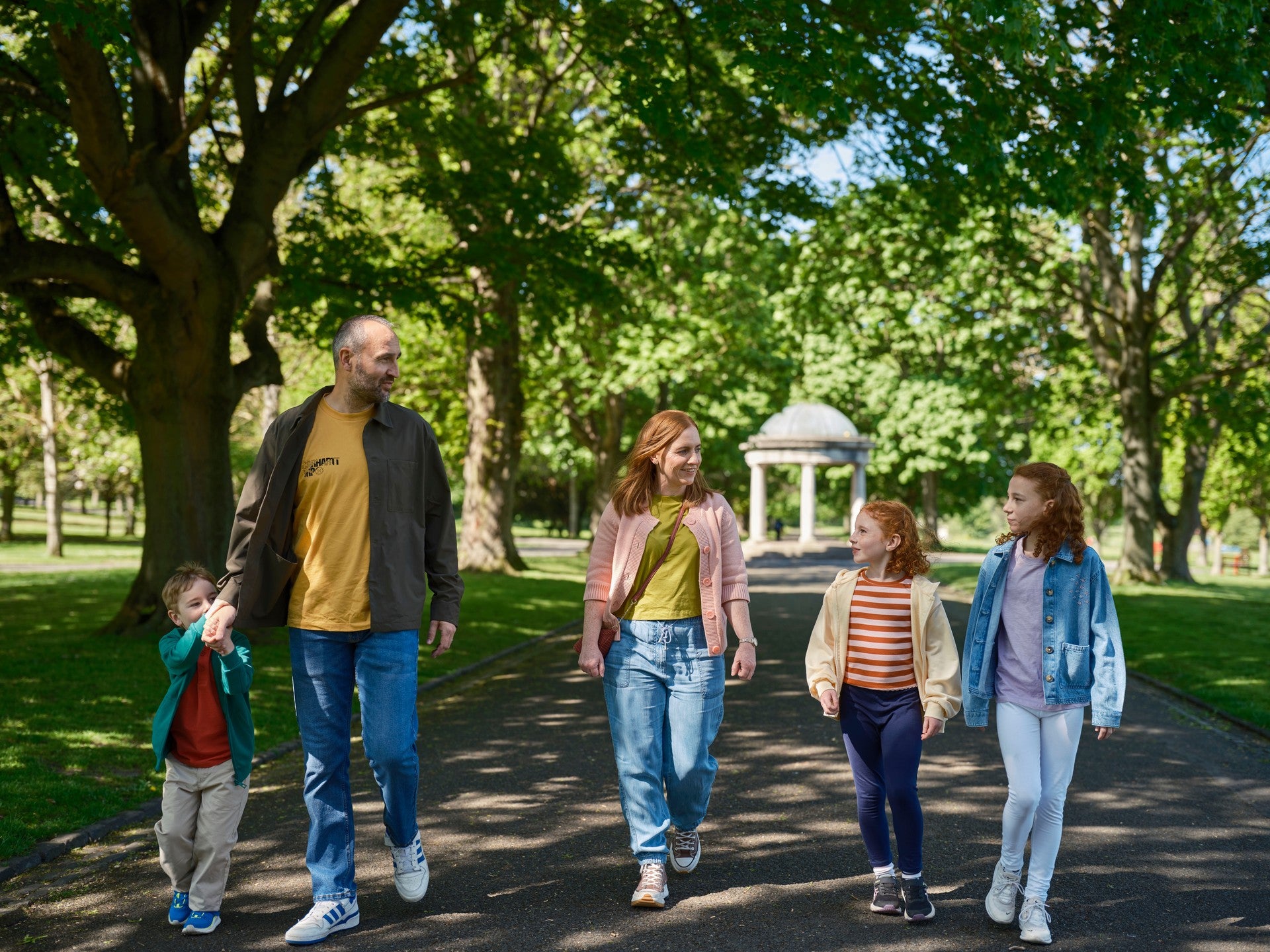 Family of five walking together along a tree-lined path in a public park on a sunny day