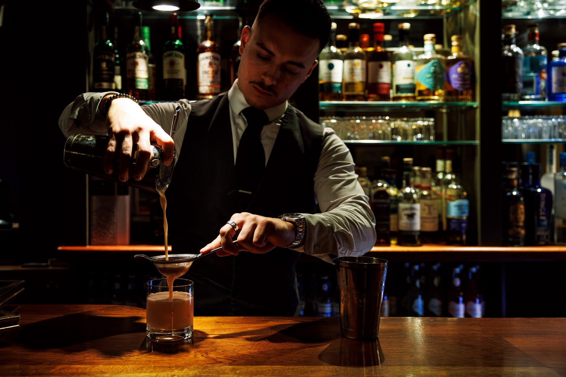 Bartender straining a cocktail into a glass at a moody, wood-panelled bar with backlit spirits shelves