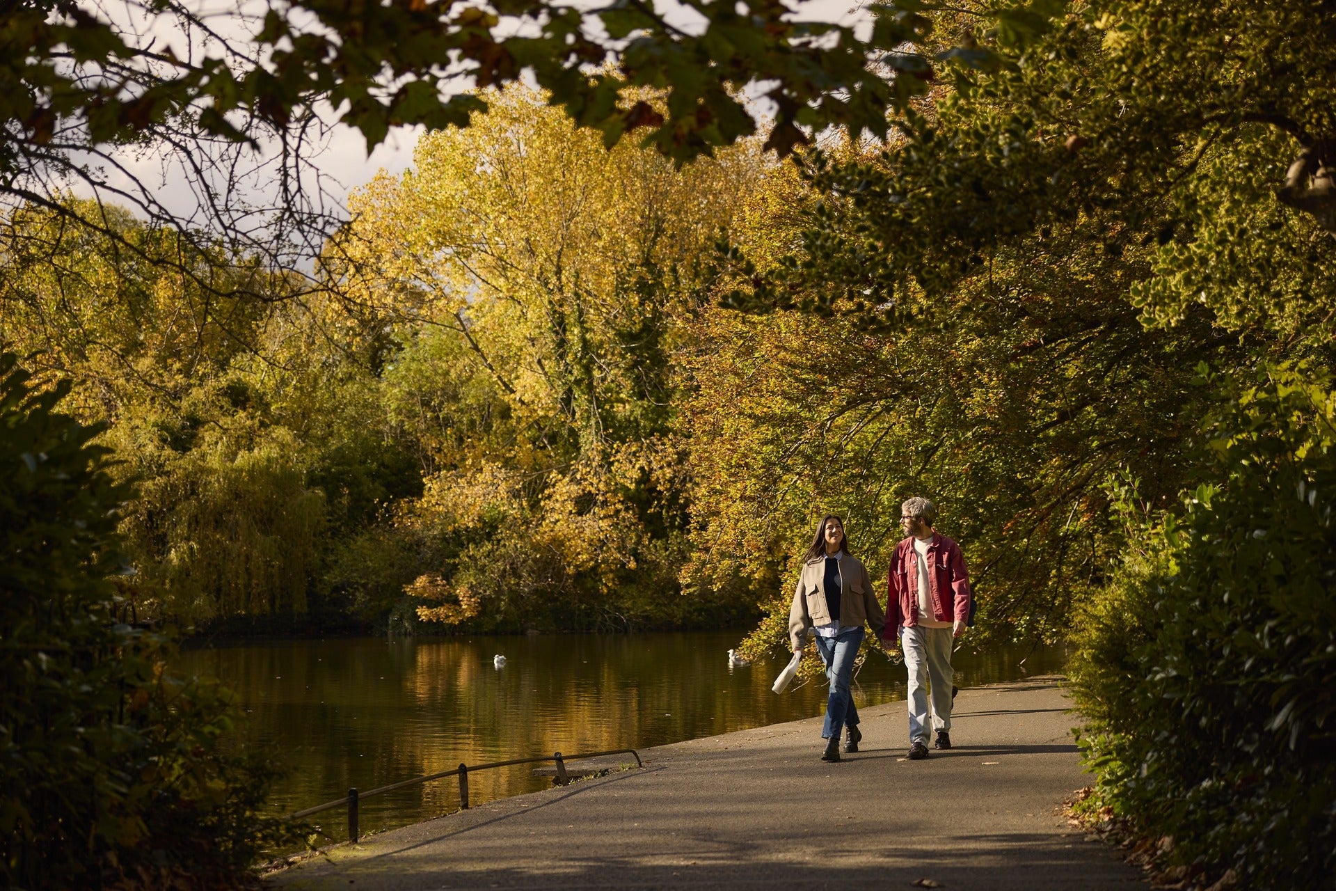 Couple walking hand-in-hand along a tree-lined path beside a lake in an autumn park with swans on the water