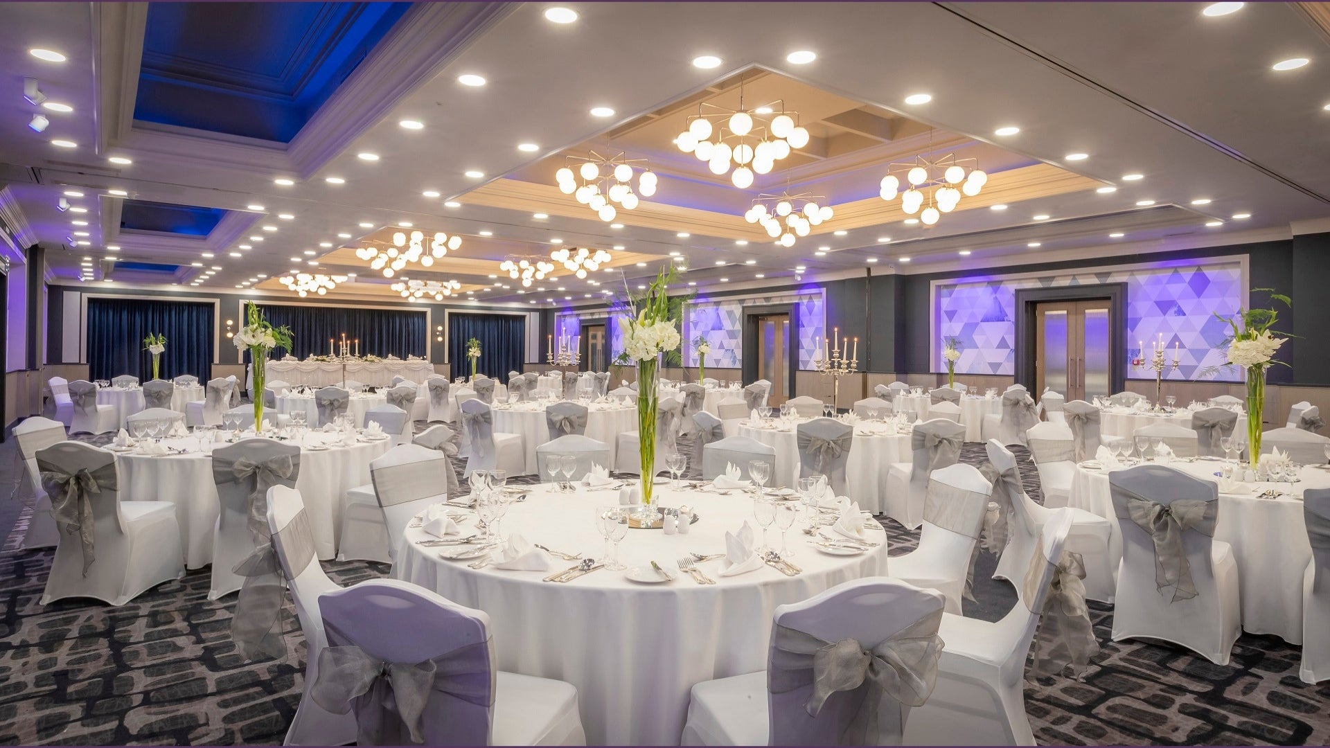 Large hotel ballroom set for a gala event, with round tables, white chair covers, grey sashes, and purple ambient lighting