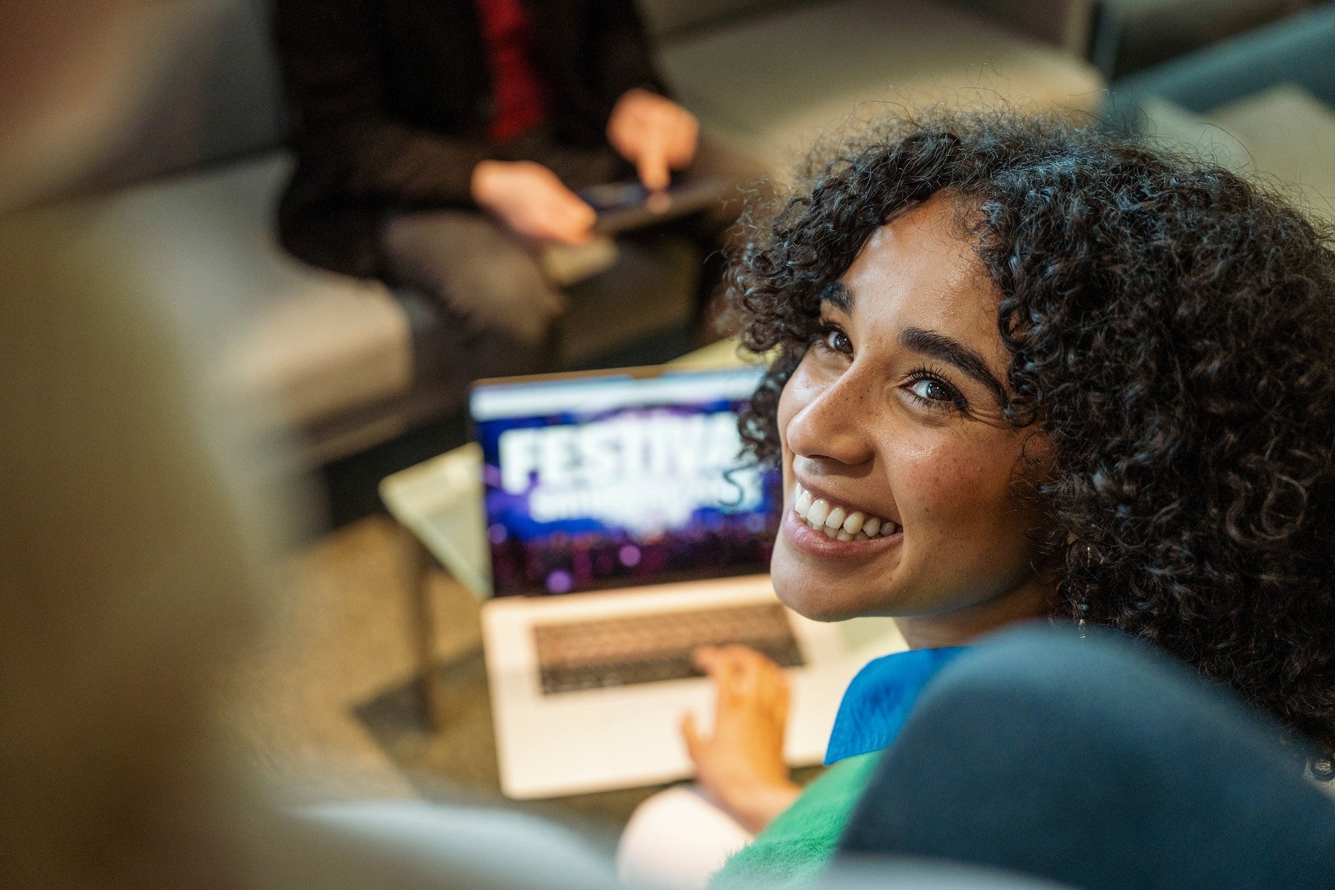 Young woman smiling while working on a laptop showing a festival website in a relaxed lounge setting