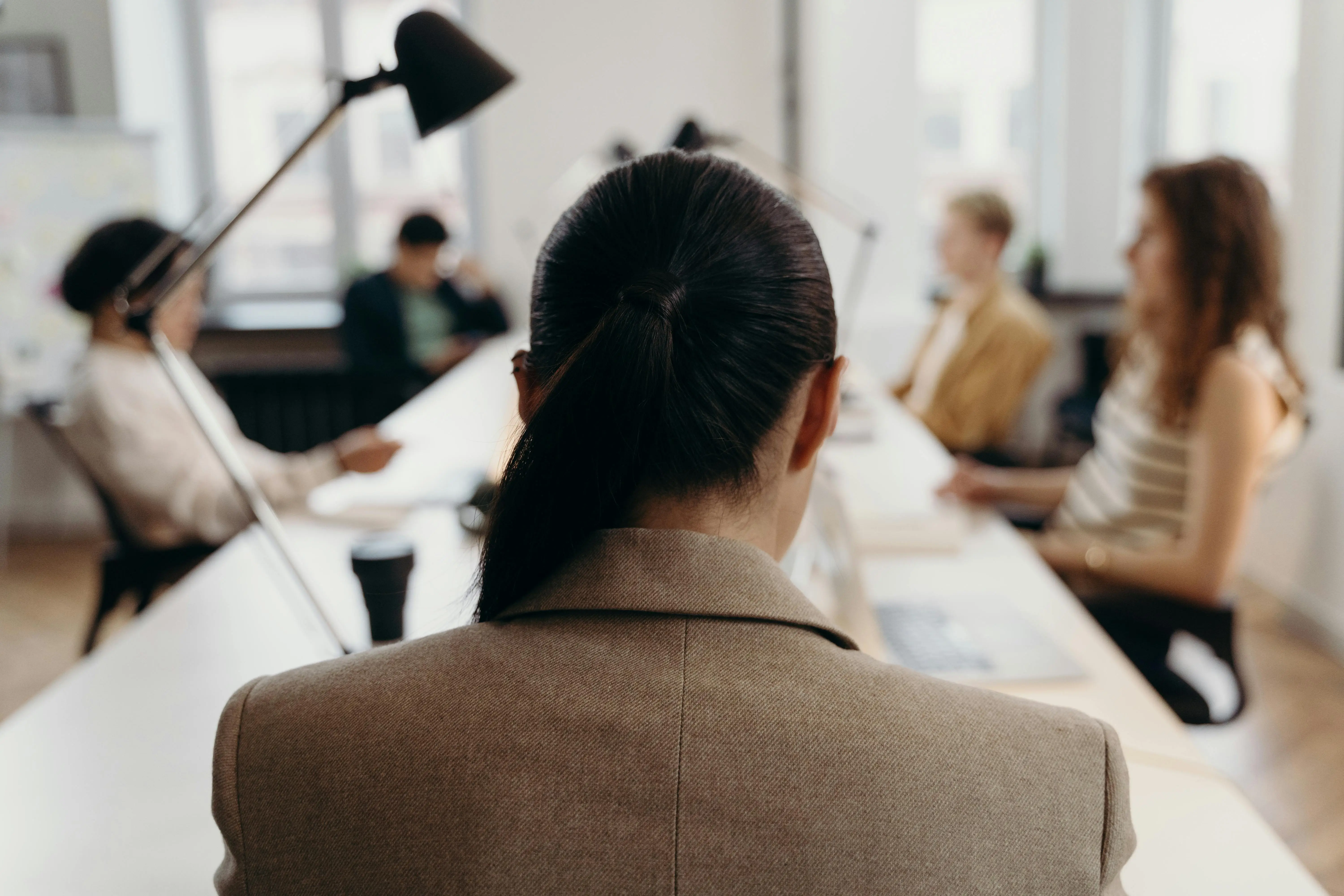 Rear view of a woman in a suit sitting at a shared desk, surrounded by colleagues in a modern office.