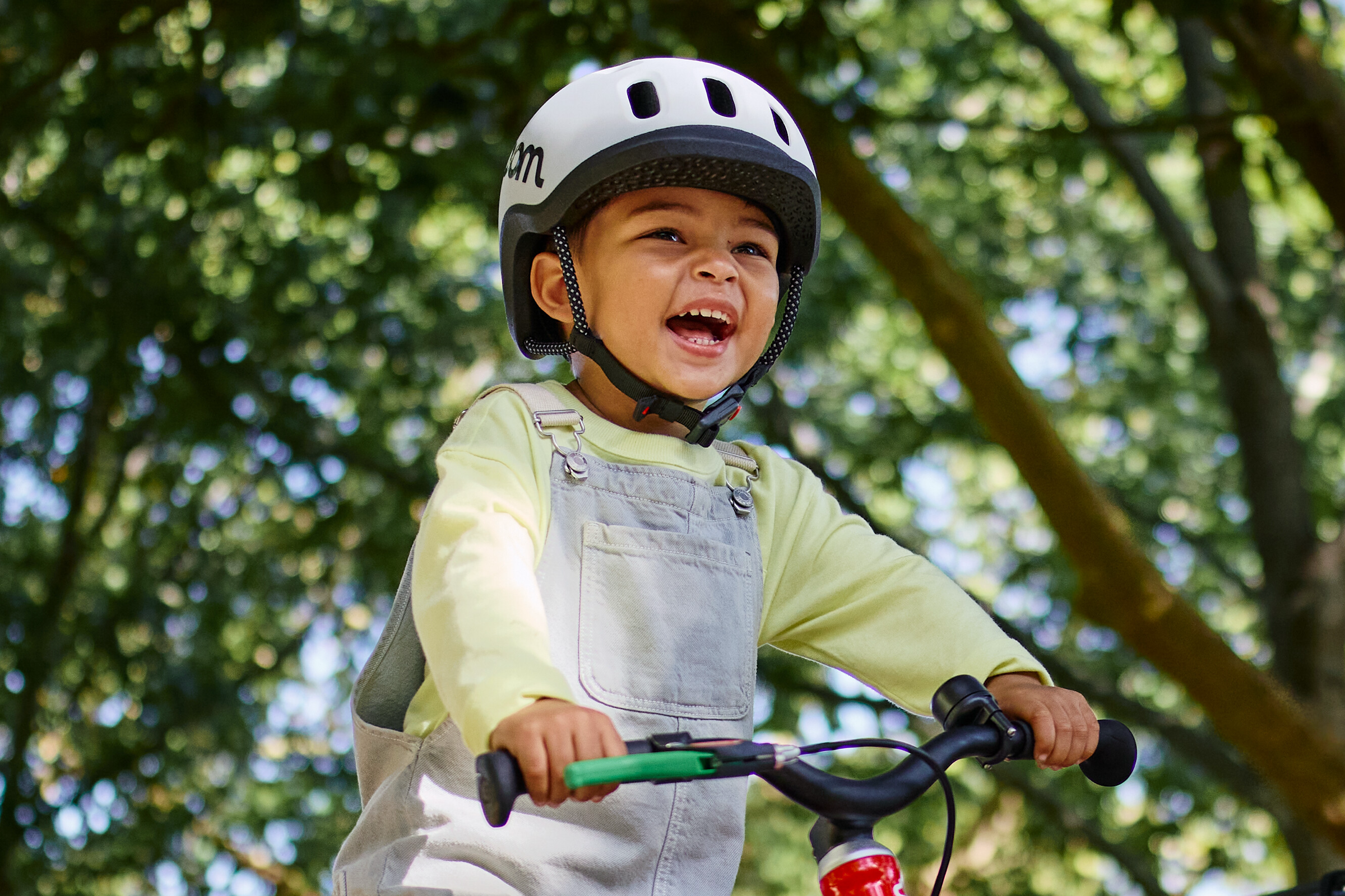 A young child wearing a READY bike helmet is holding the handlebars of a red woom GO 1 balance bike in front of some trees.