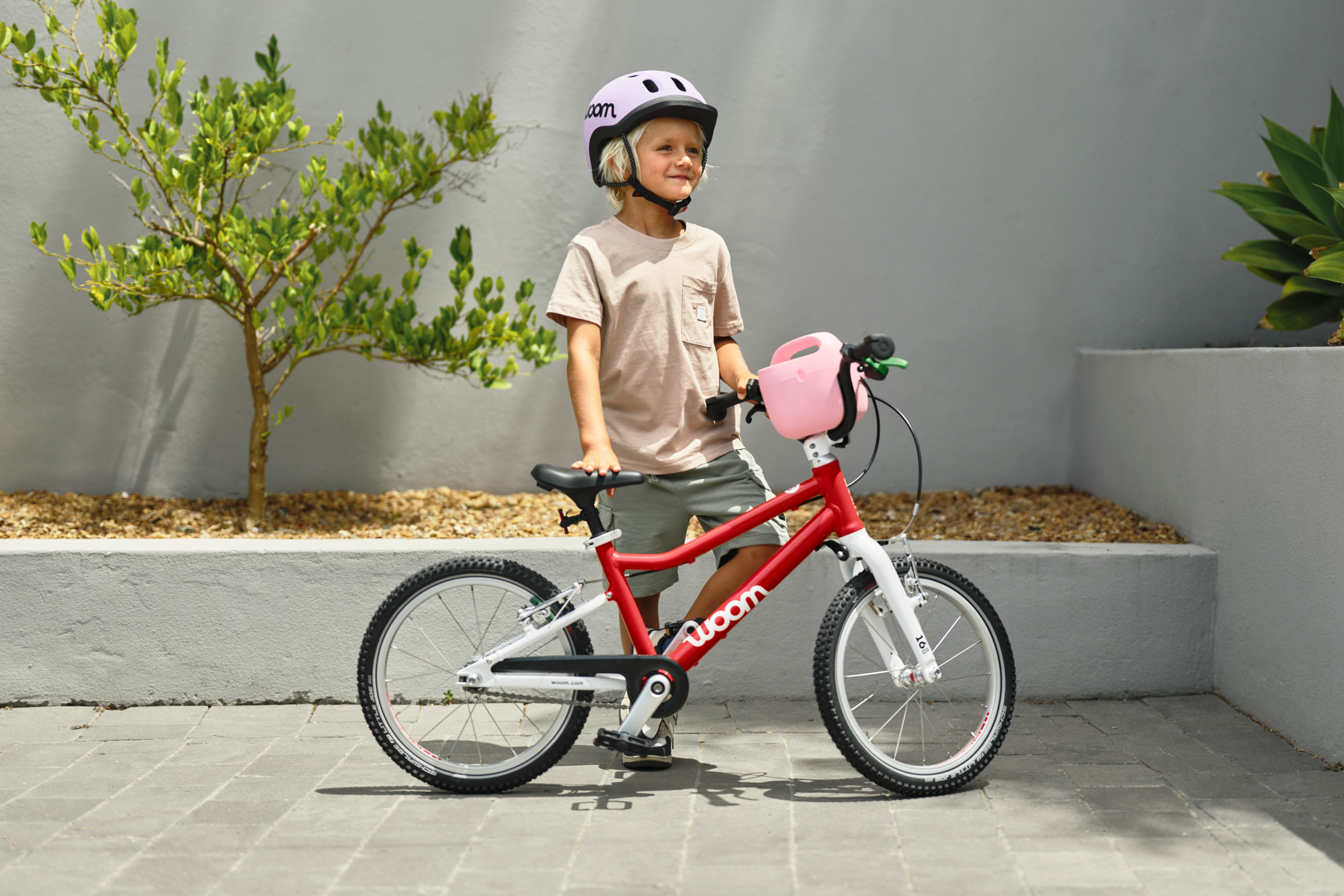 A child in a lilac READY helmet stands behind a red woom GO 3 with a pink POP bike basket on the handlebars