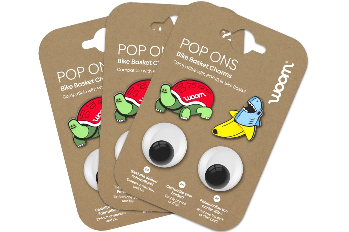 Paquet de pin’s POP ONS pour panier vélo avec quatre pin’s : deux gros yeux mobiles, une tortue dont la carapace a été remplacée par un casque woom rouge, et un pin’s mi-banane, mi-requin.