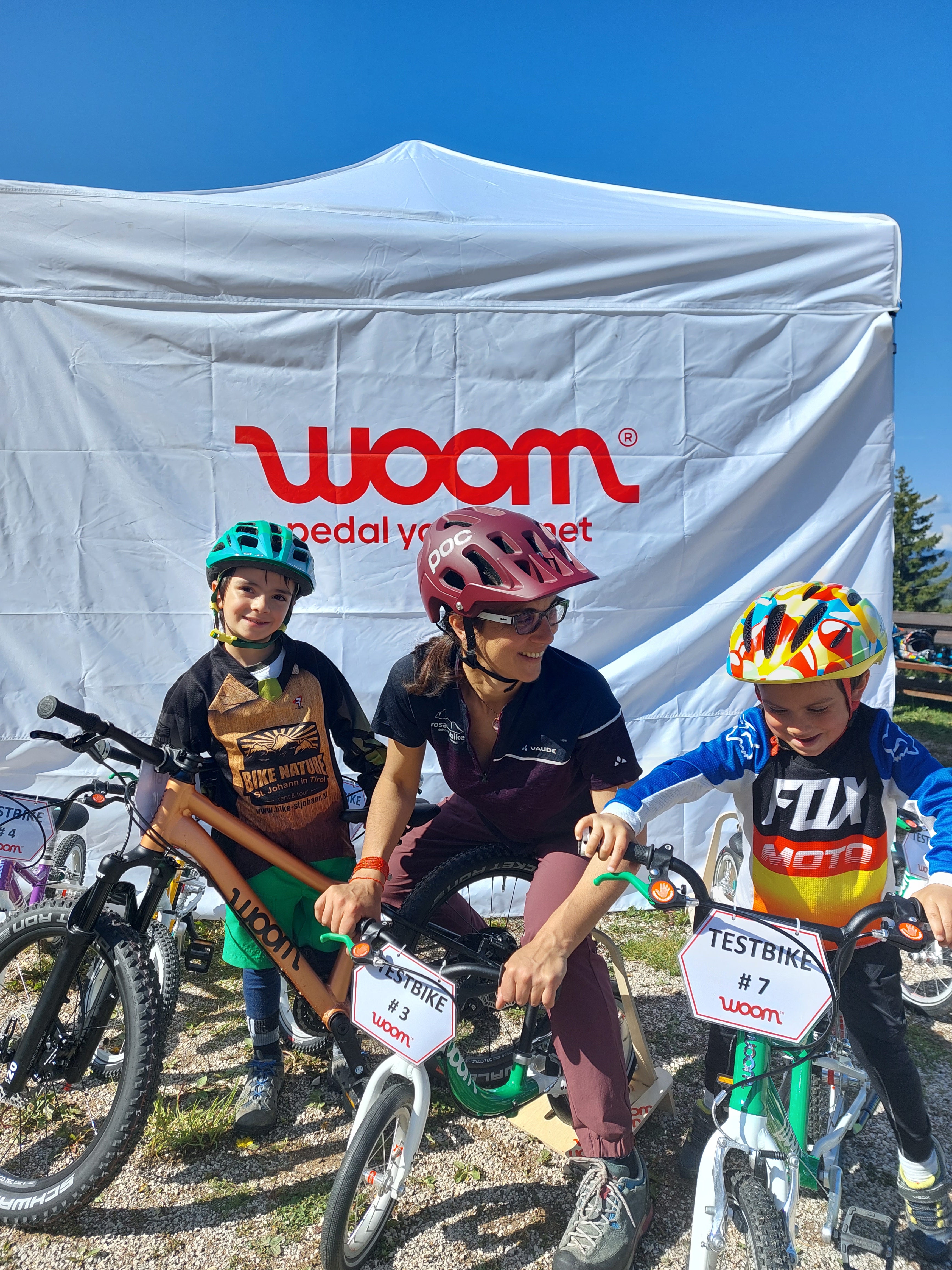 A mother poses on a woom balance bike, with her two sons either side also on woom bikes.