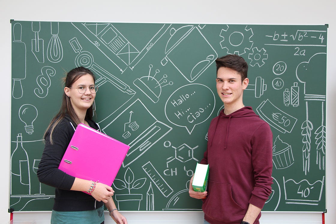 Schülerin mit einer magenta Mappe und Schüler mit Wörterbuch in der Hand vor grüner Schultafel mit Kreidezeichnungen zu verschiedenen Unterrichtfächer